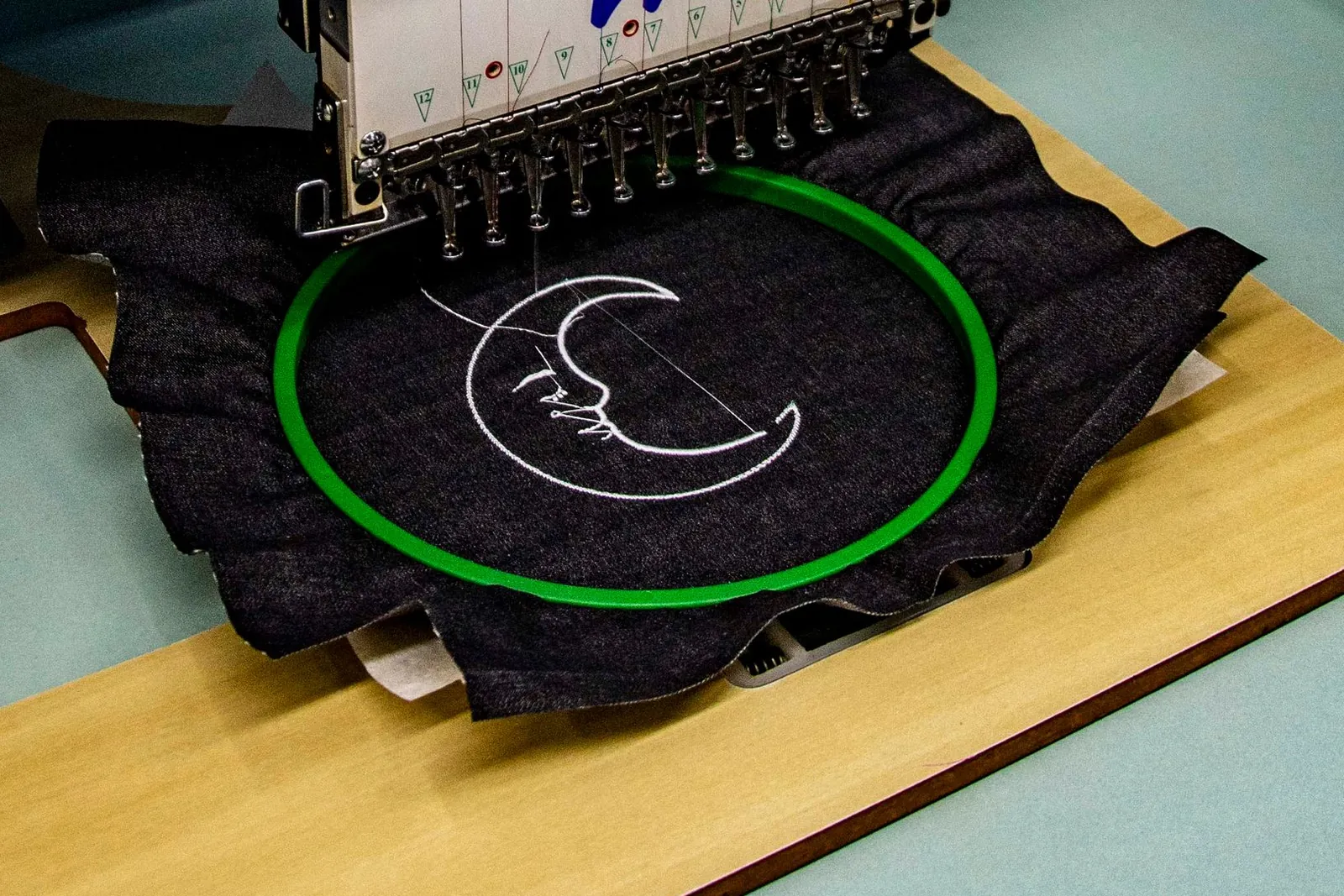An embroidery machine stitching a moon design onto a dark garment held in a green hoop