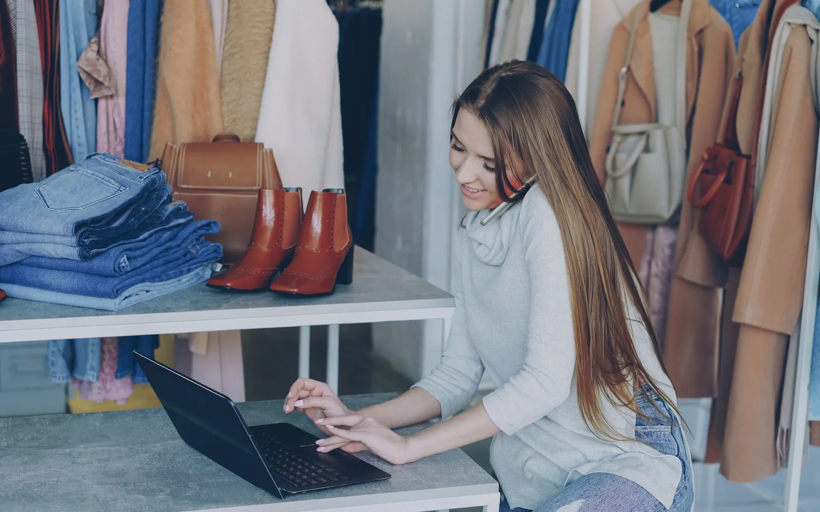 A woman in a clothing boutique on the phone while typing on a laptop, surrounded by folded jeans, boots and coats