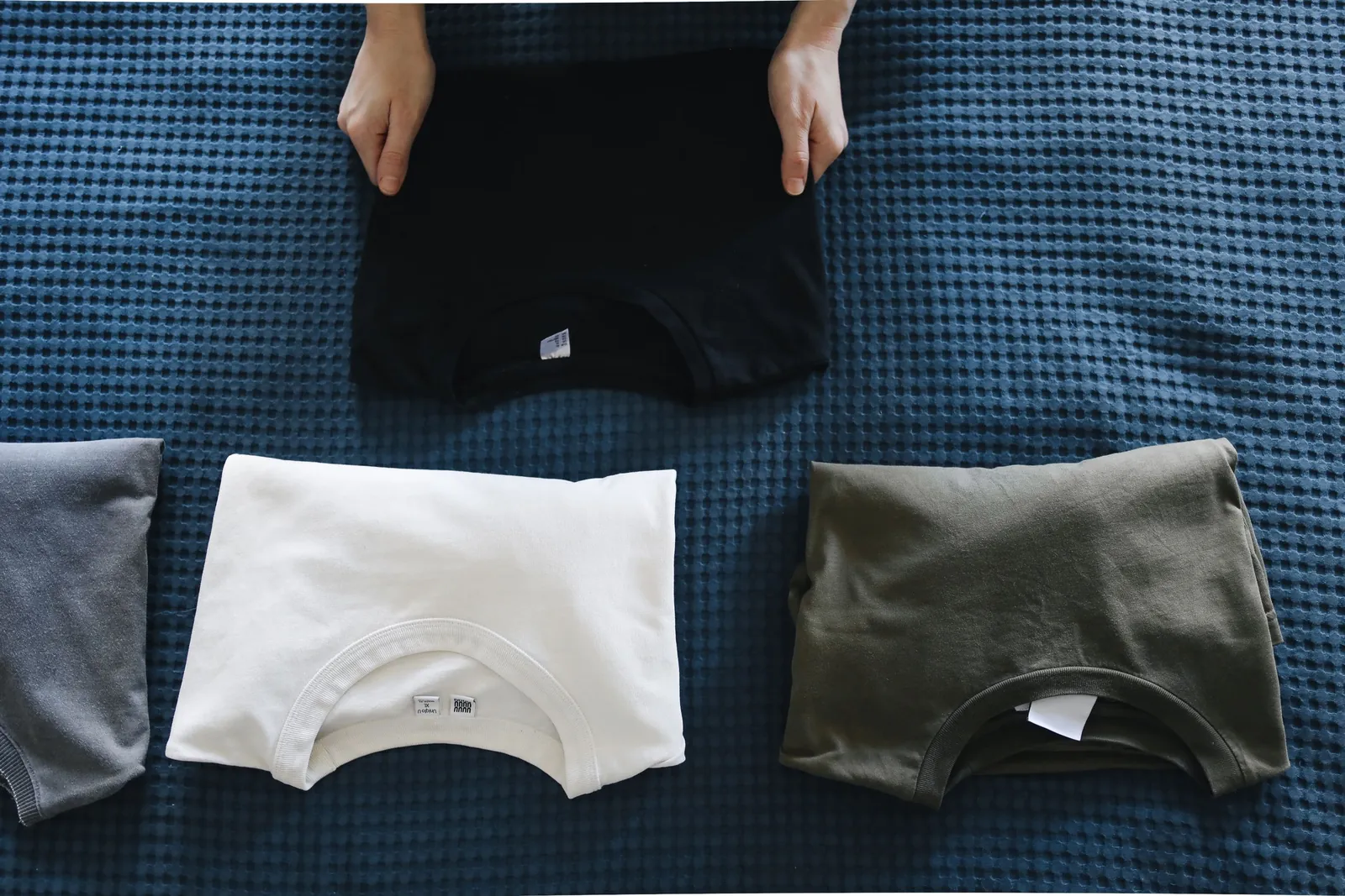 Folded T-shirts in black, white and olive laid out on a bed