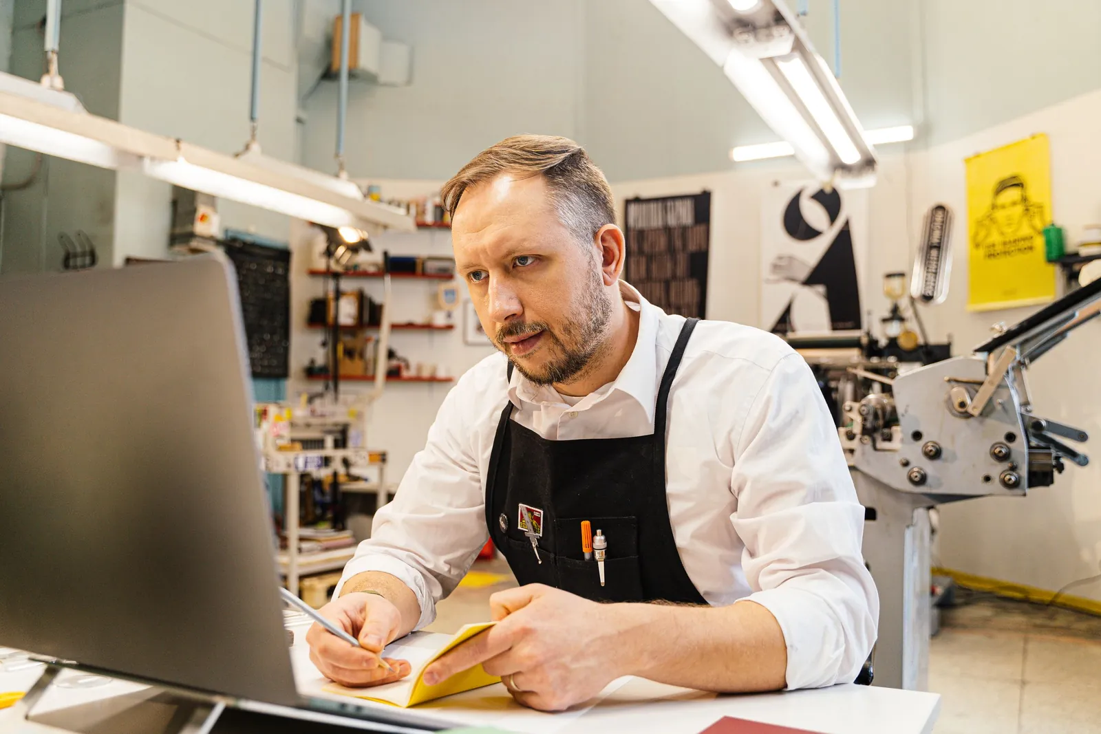 A print shop owner at his desk, working through an order on a laptop with notes beside him