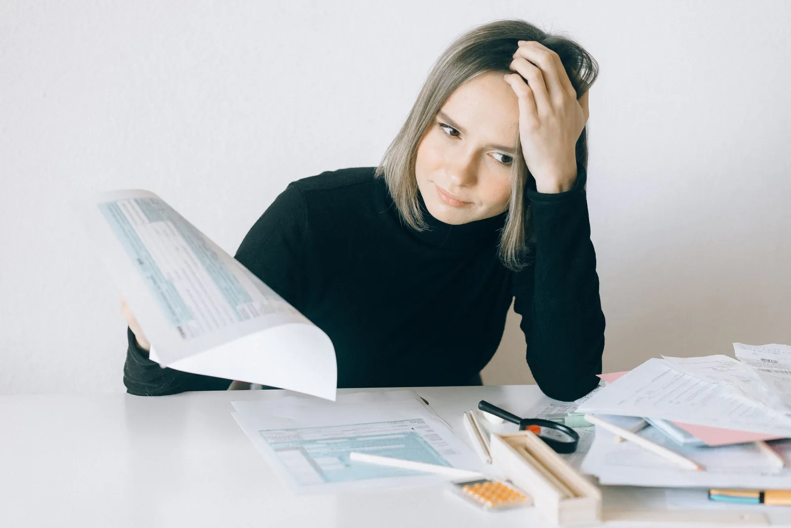 A woman at a desk holding her head while reading through a stack of printed paperwork