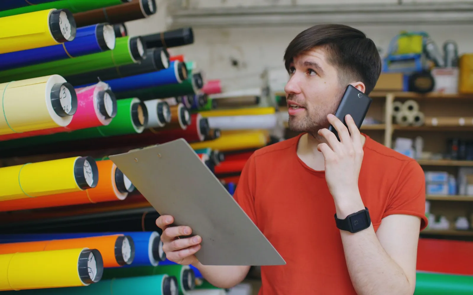 A warehouse worker on the phone holding a clipboard, in front of shelves of colorful vinyl rolls