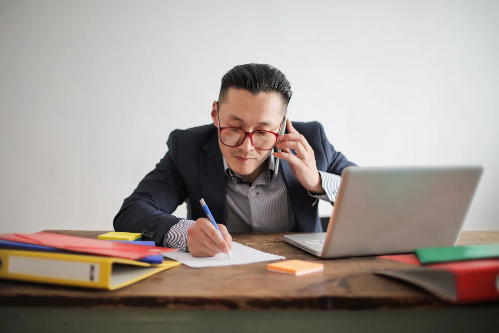 A man on a phone call writing notes at a desk with a laptop and colored binders